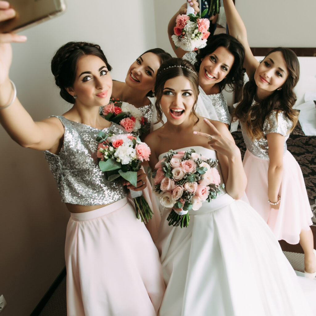 Bride and 4 bridesmaids holding flowers and posing for a selfie.