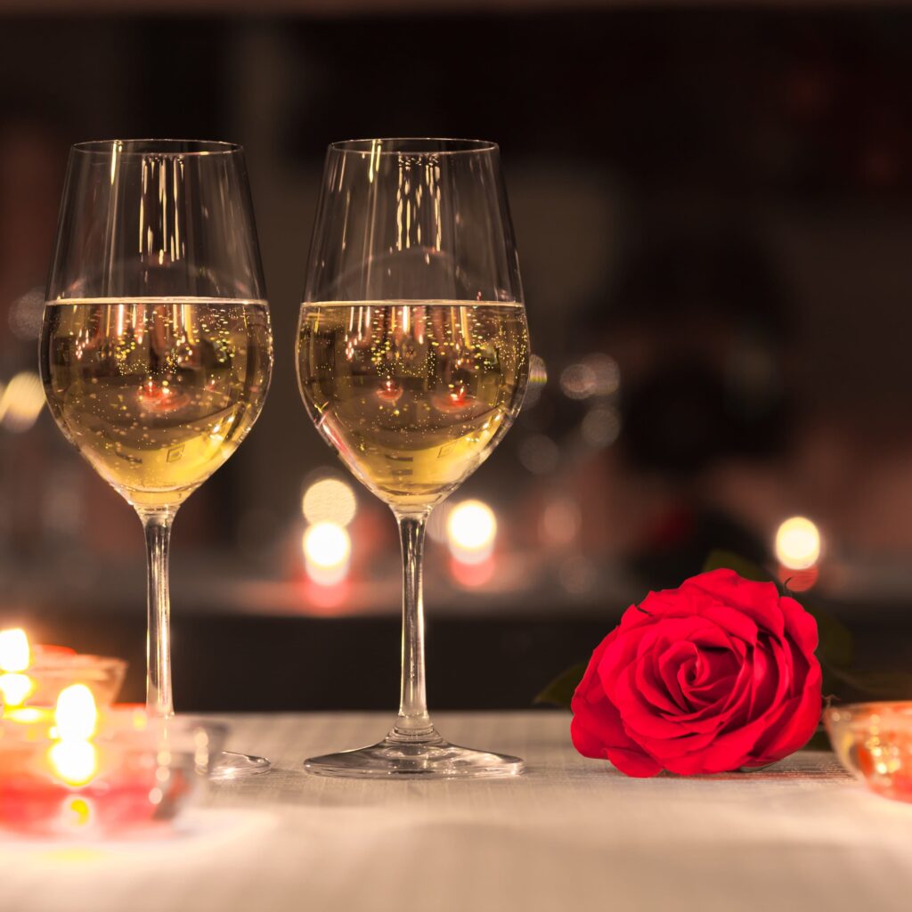 Two glasses of white wine on restaurant table with red rose and soft lighting.