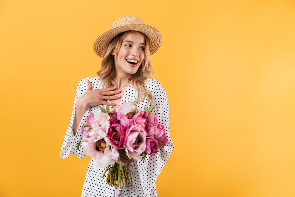 Girl wearing straw hat and nice dress, holding pink flowers on yellow background.